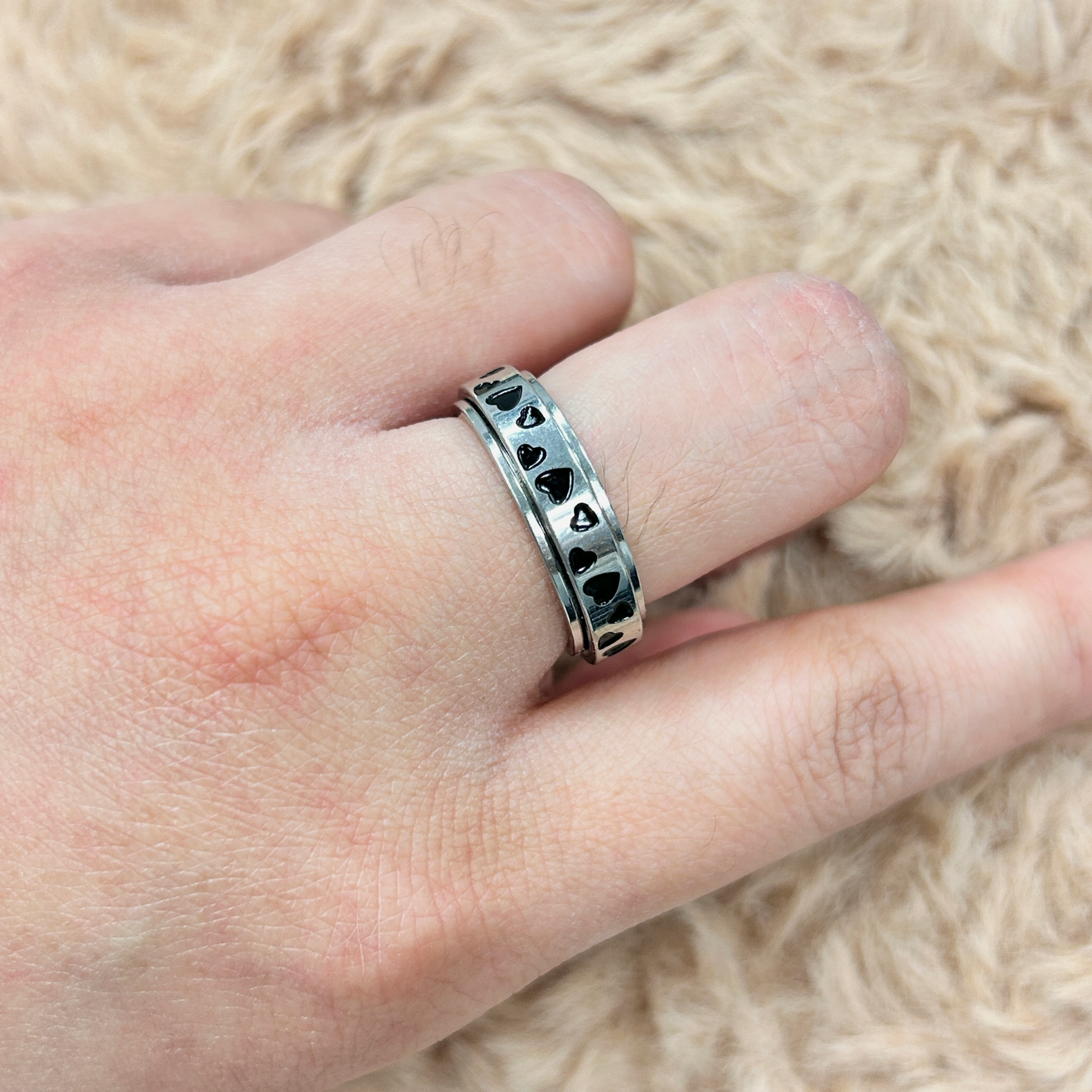 Close-up of a finger wearing a silver ring with repeating heart shapes, set against a soft beige fur background.