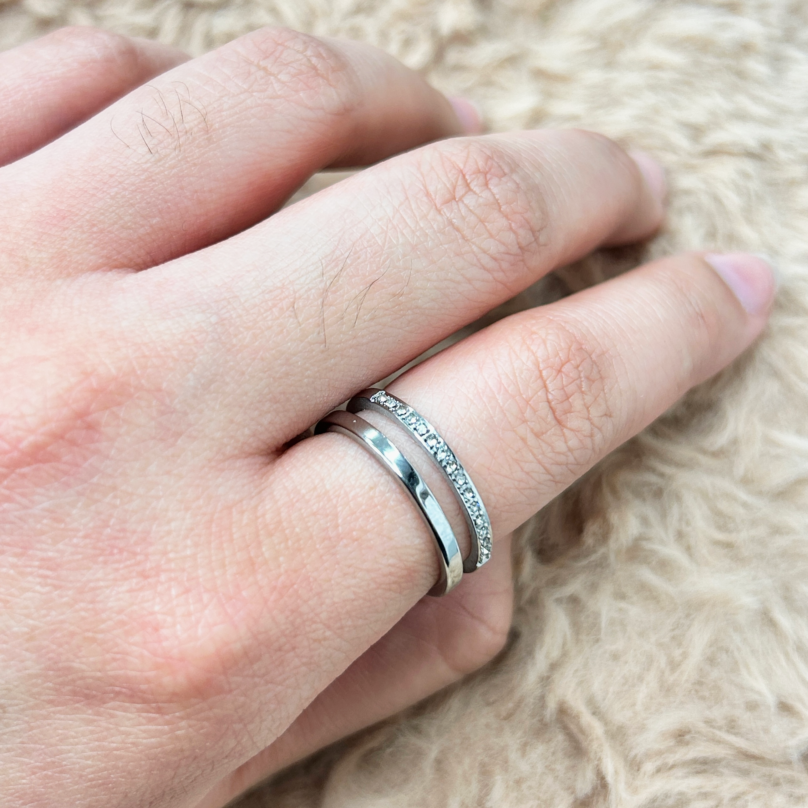 Close-up of a hand with two silver rings on the ring finger, one plain band and one with small stones, resting on a soft beige surface.