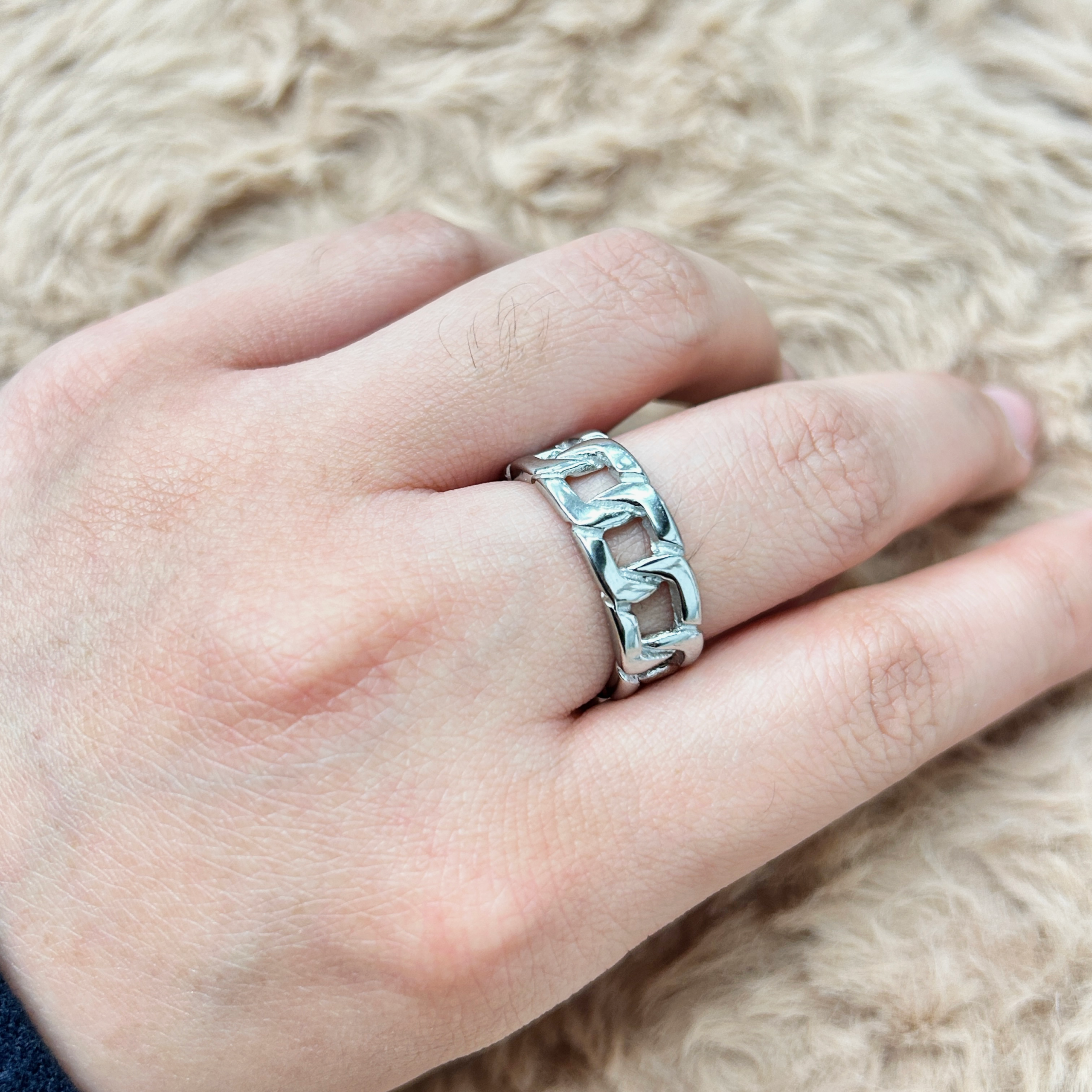 Close-up of a silver ring on a finger, with a textured, chain-link design, against a beige fuzzy background.