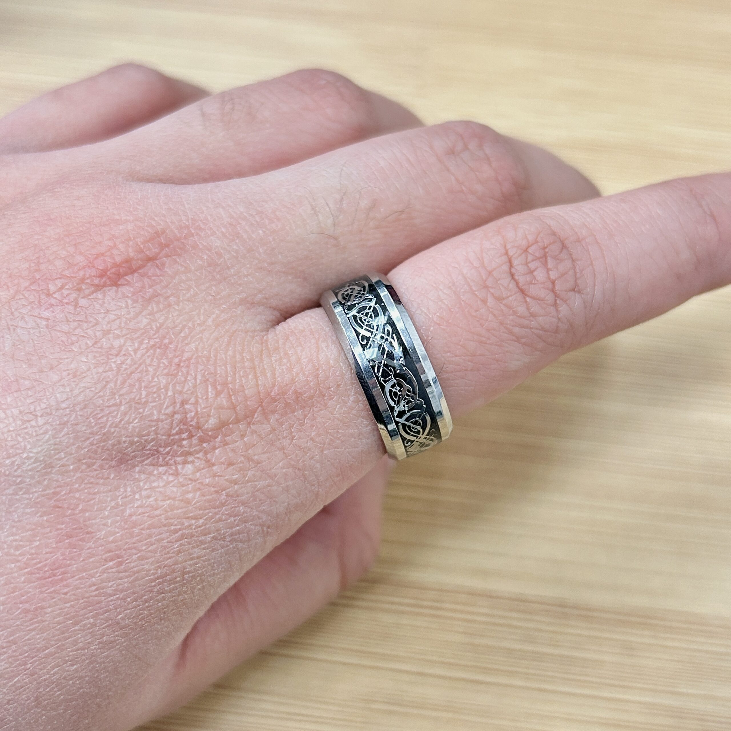 Close-up of a finger wearing a silver ring with an intricate black vine pattern on a light wooden background