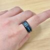 Close-up of a finger wearing a gray metal ring with a blue inlay on the skin, wooden table in the background.