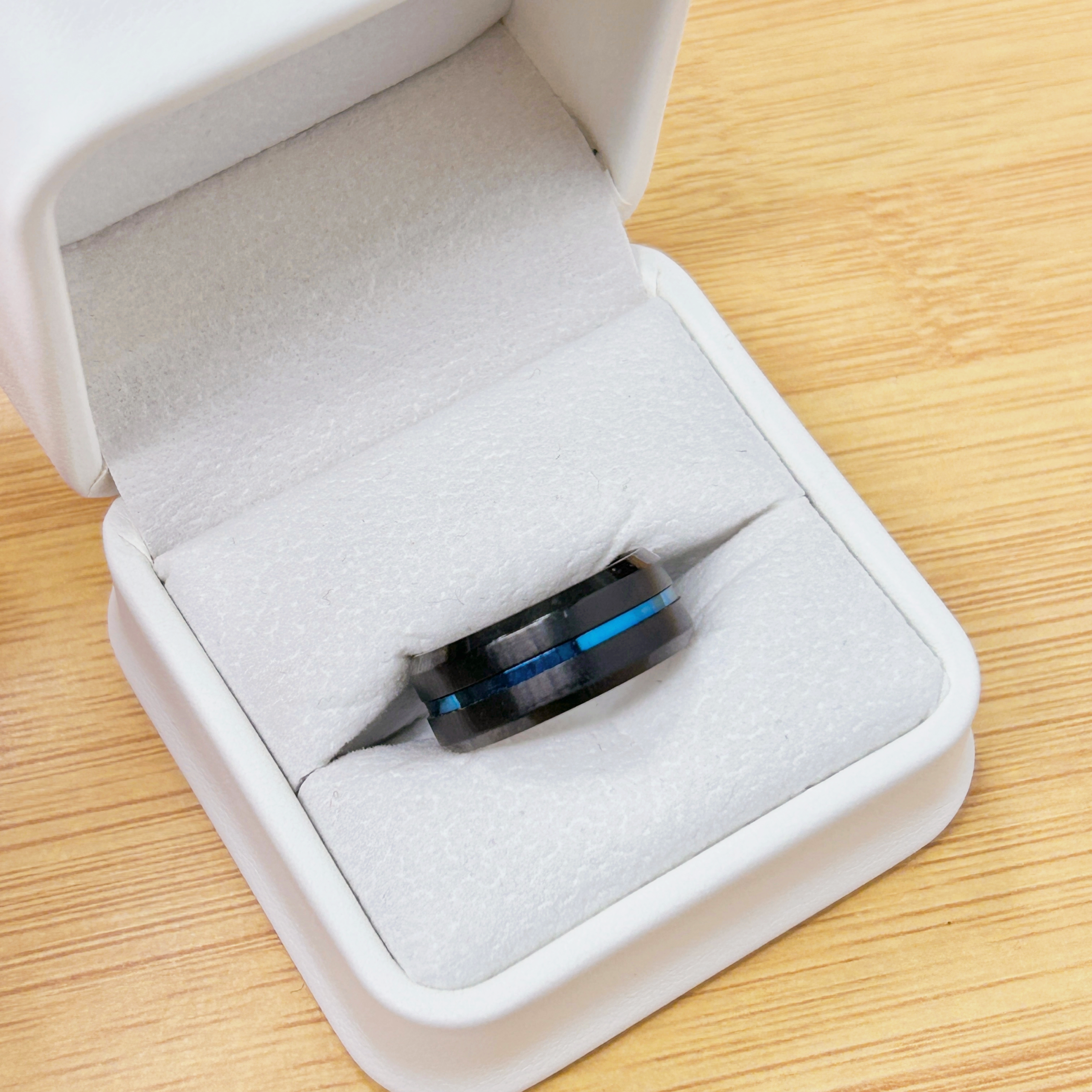 Open white foam-lined jewelry box on a wooden table with a blue-and-black ring or bracelet inside