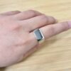 Close-up of a silver rectangular ring on a finger resting on a light wooden surface.