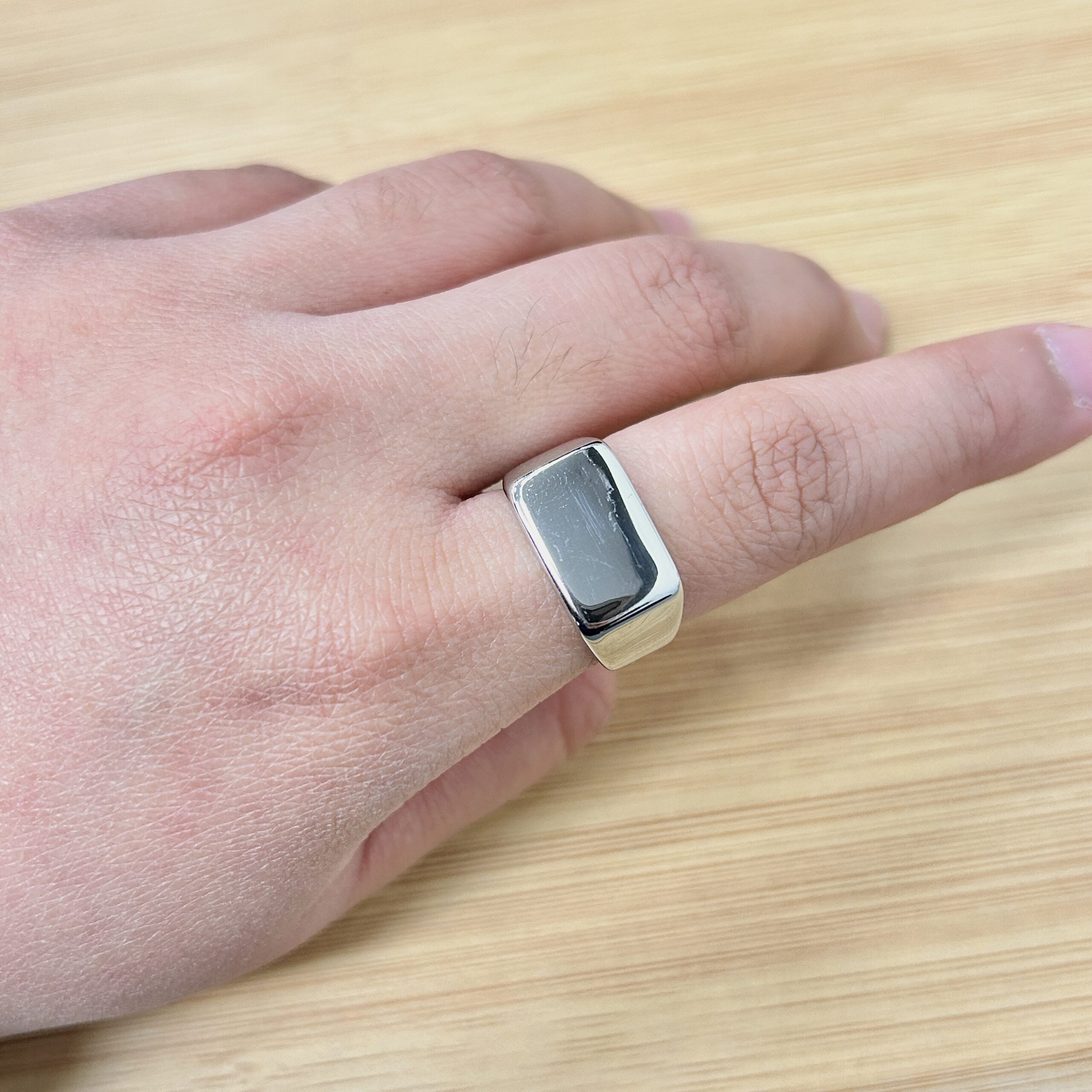 Bold Silver Square Shiny Statement Ring - Eriy Gift Close-up of a silver rectangular ring on a finger resting on a light wooden surface.