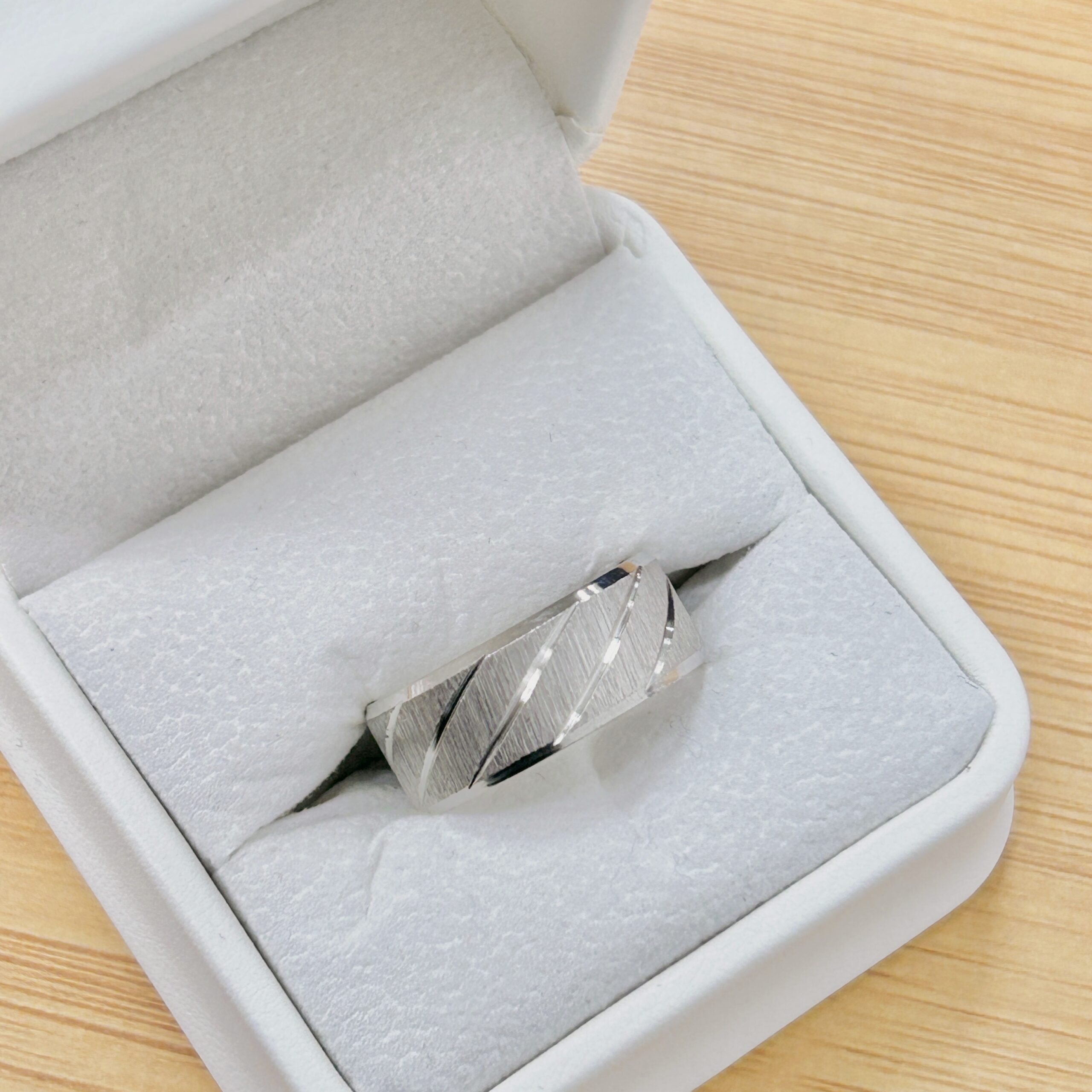 Close-up of a silver ring resting in a light gray jewelry box on a wooden table surface, a soft velvet interior visible around it.