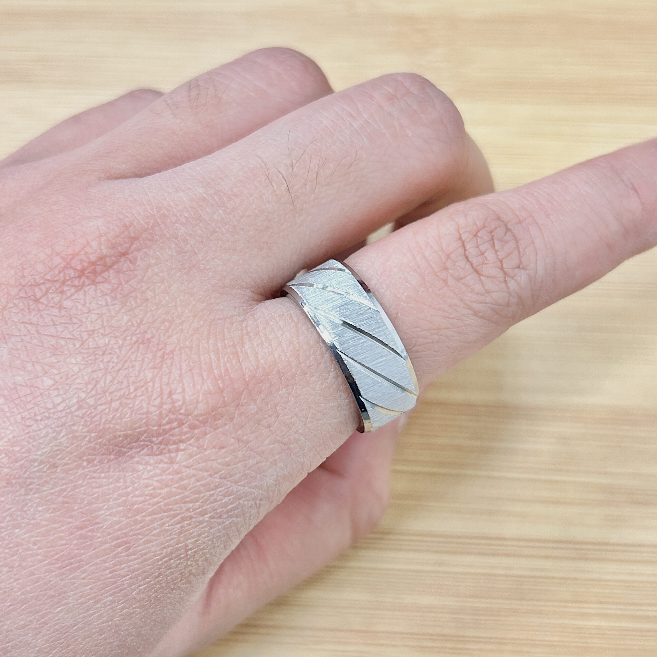 Close-up of a faceted silver ring with diagonal stripes on a finger, against a light wood background.