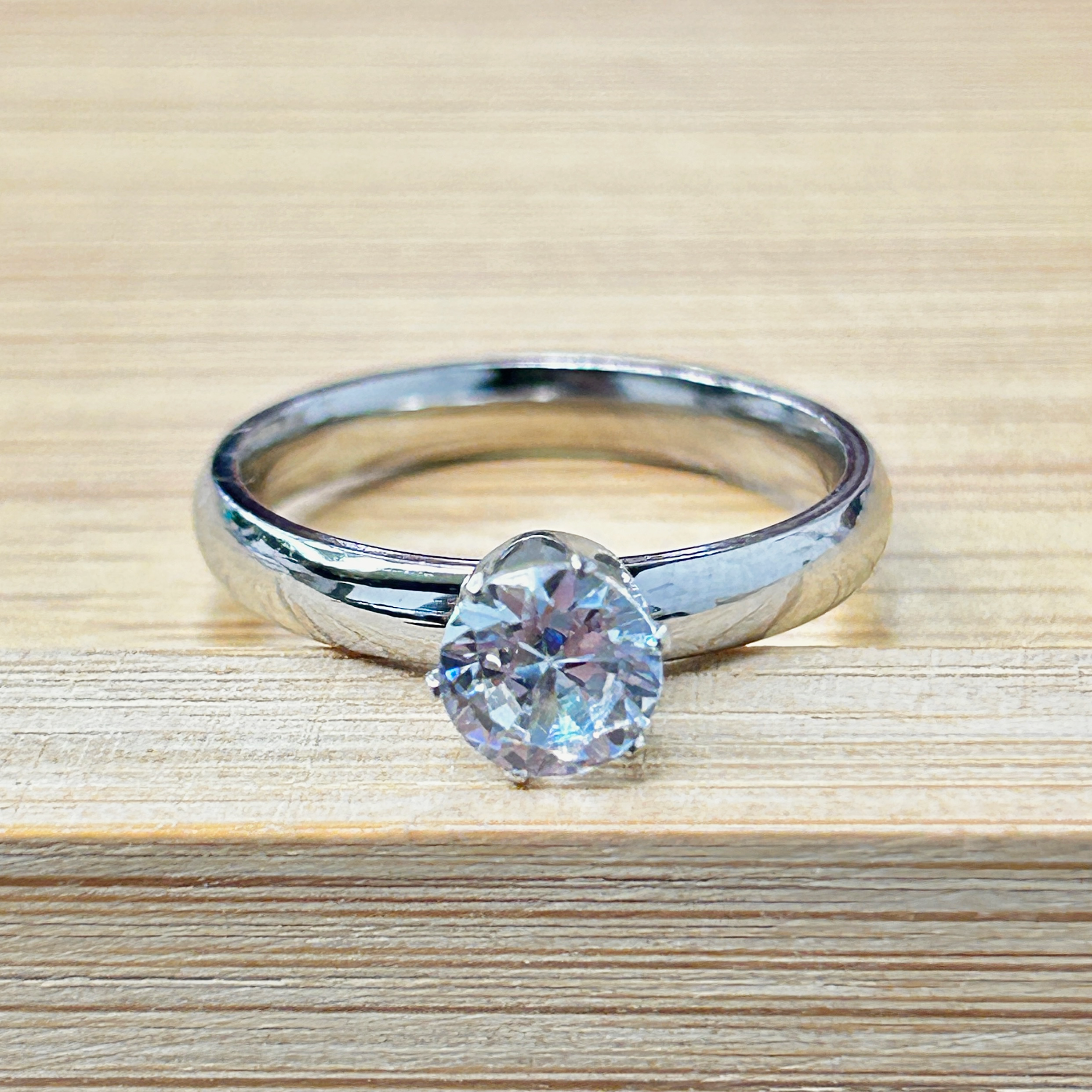 Classic Silver Solitaire Promise Ring picture2 - Eriy Gift Close-up of a diamond engagement ring with a large faceted diamond on a silver band, resting on a wooden surface, with a second plain band nearby.