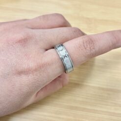 Close-up of a silver patterned ring on a finger, resting on a light wooden surface.
