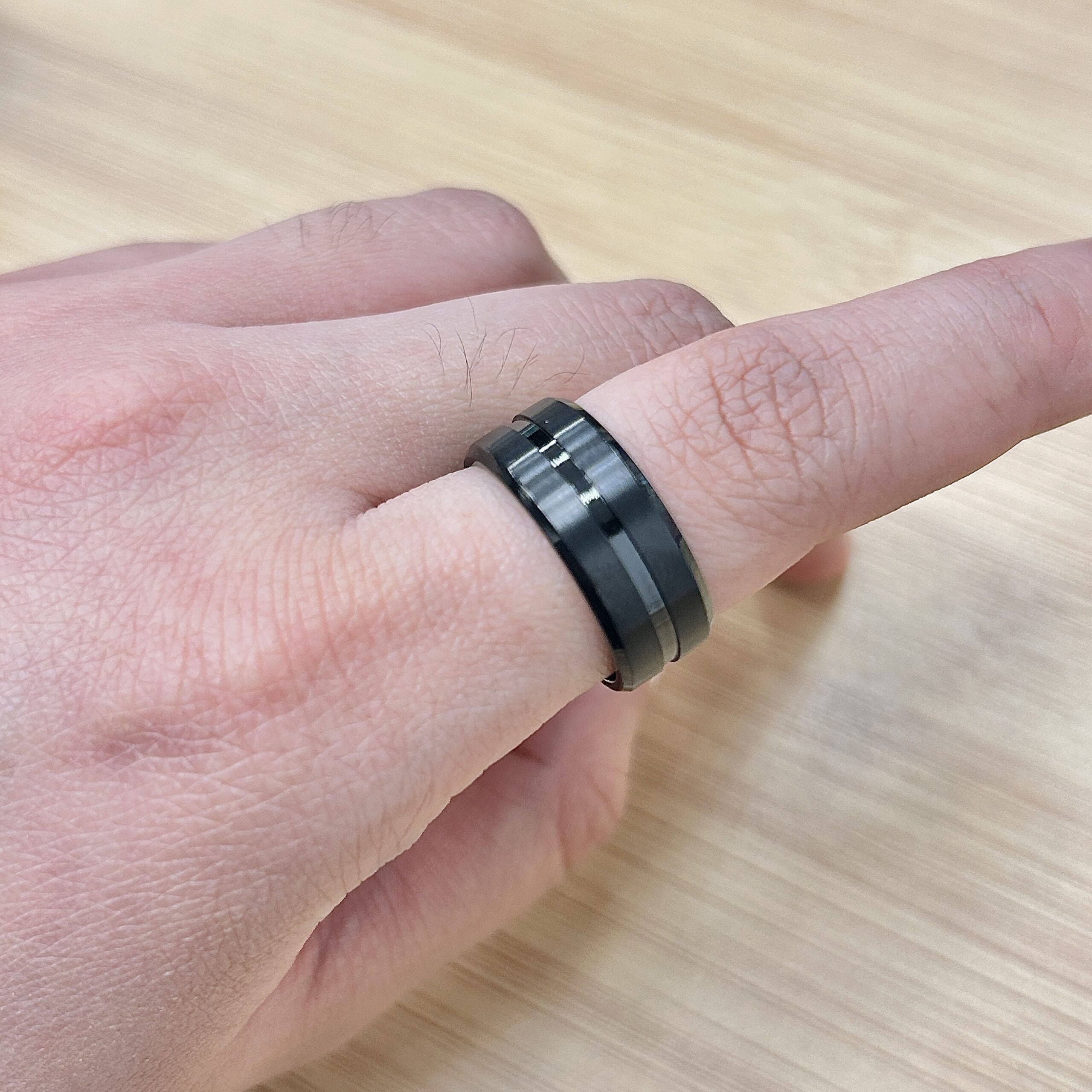 Matte Black Center Groove Ring - Eriy Gift Close-up of a finger wearing a dark, segmented metal ring on a light wooden surface.