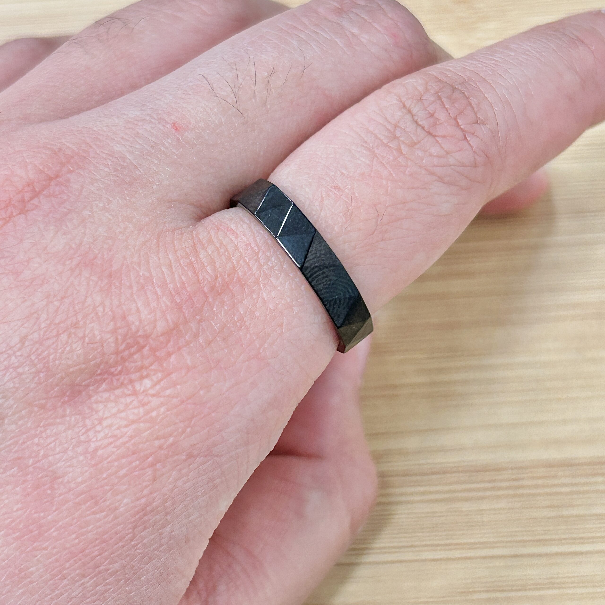 Close-up of a hand with a faceted black metal ring on the finger, against a wooden surface.
