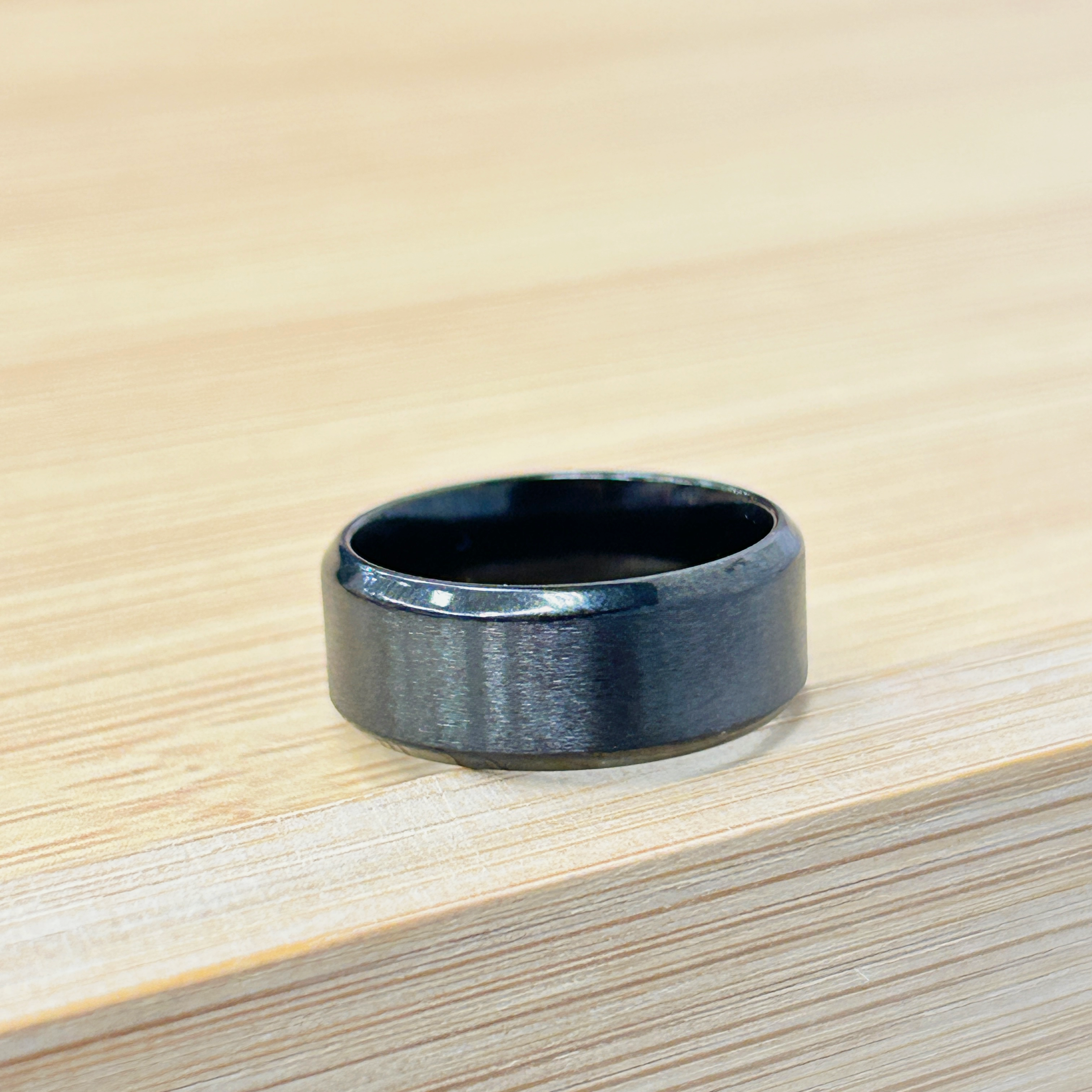 Close-up of a dark metal ring lying on a light wooden table, with a visible rough outer edge and interior hollow space.