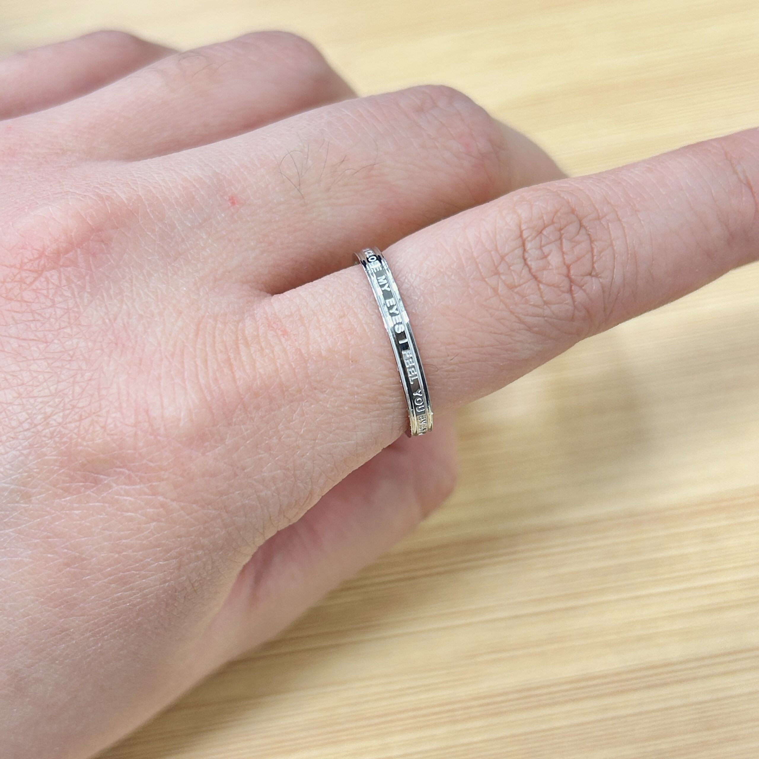 Minimalist Silver Engraved Quote Band Ring - Eriy Gift Close-up of a hand wearing a silver ring with engraved letters along the band on the finger edge, near a light wooden surface.