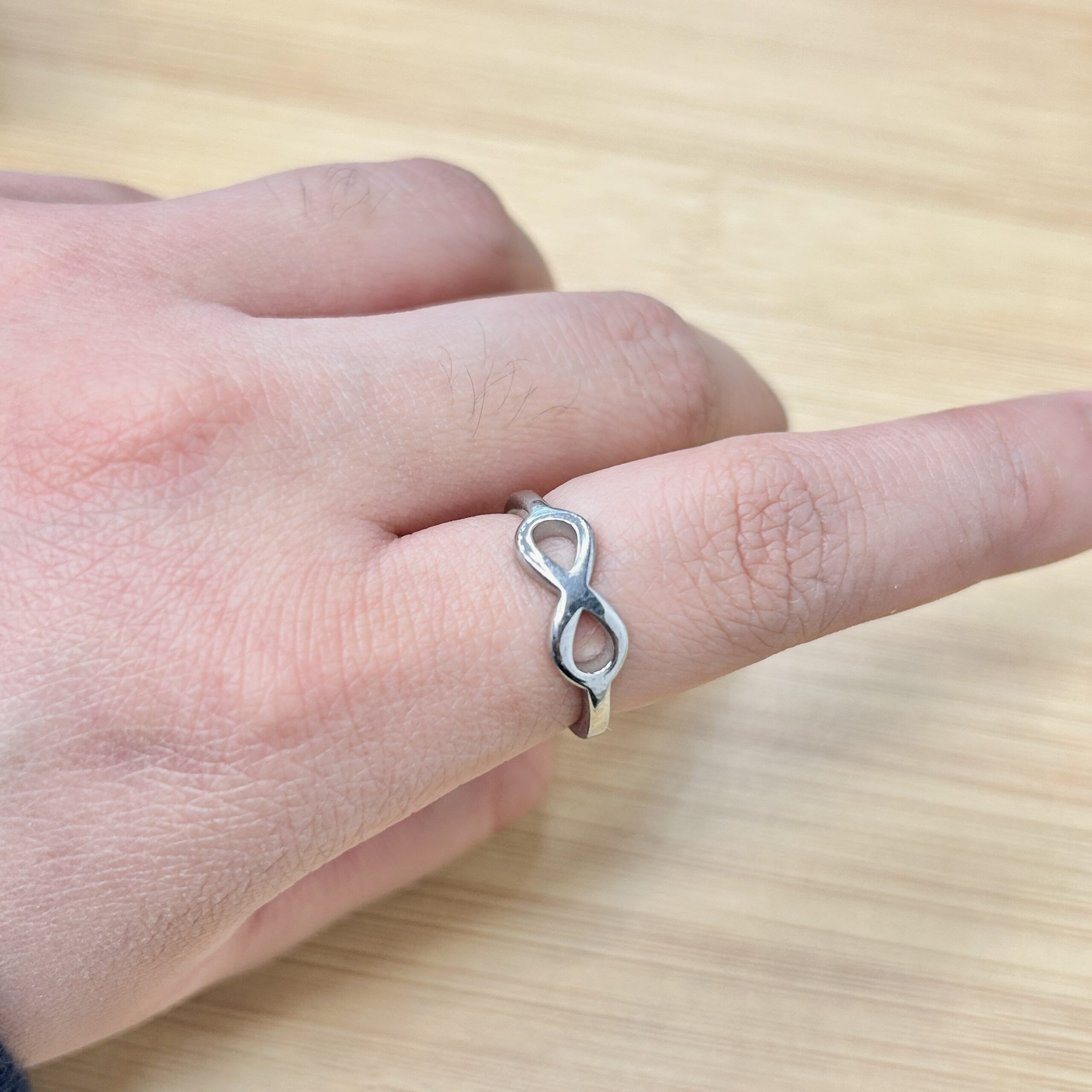 Close-up of a silver infinity-symbol ring worn on a finger, with a light wood surface in the background.