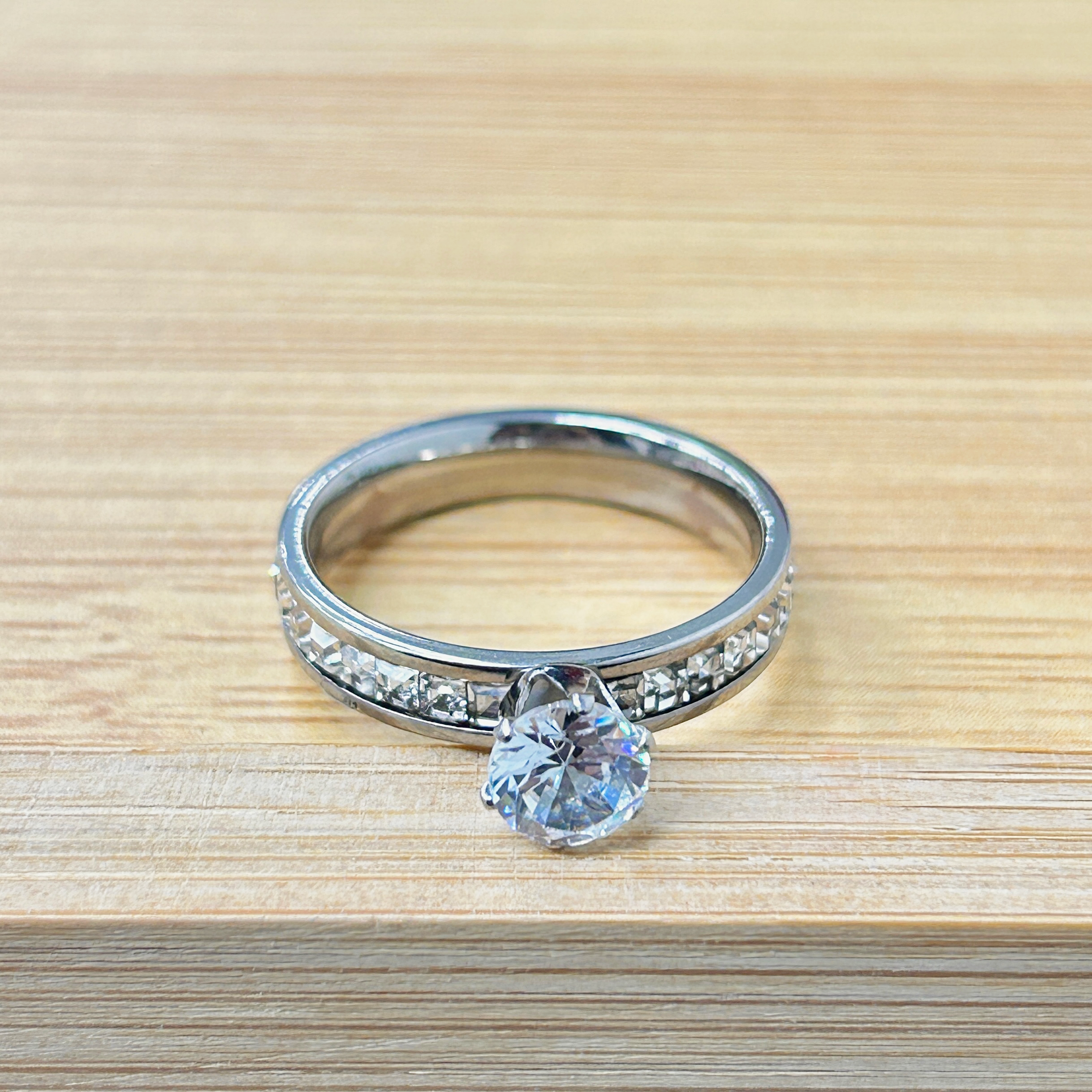 Silver Channel-Set Side Stone Solitaire Ring picture2 - Eriy Gift Close-up of a silver ring with a large clear diamond on a light wooden surface.