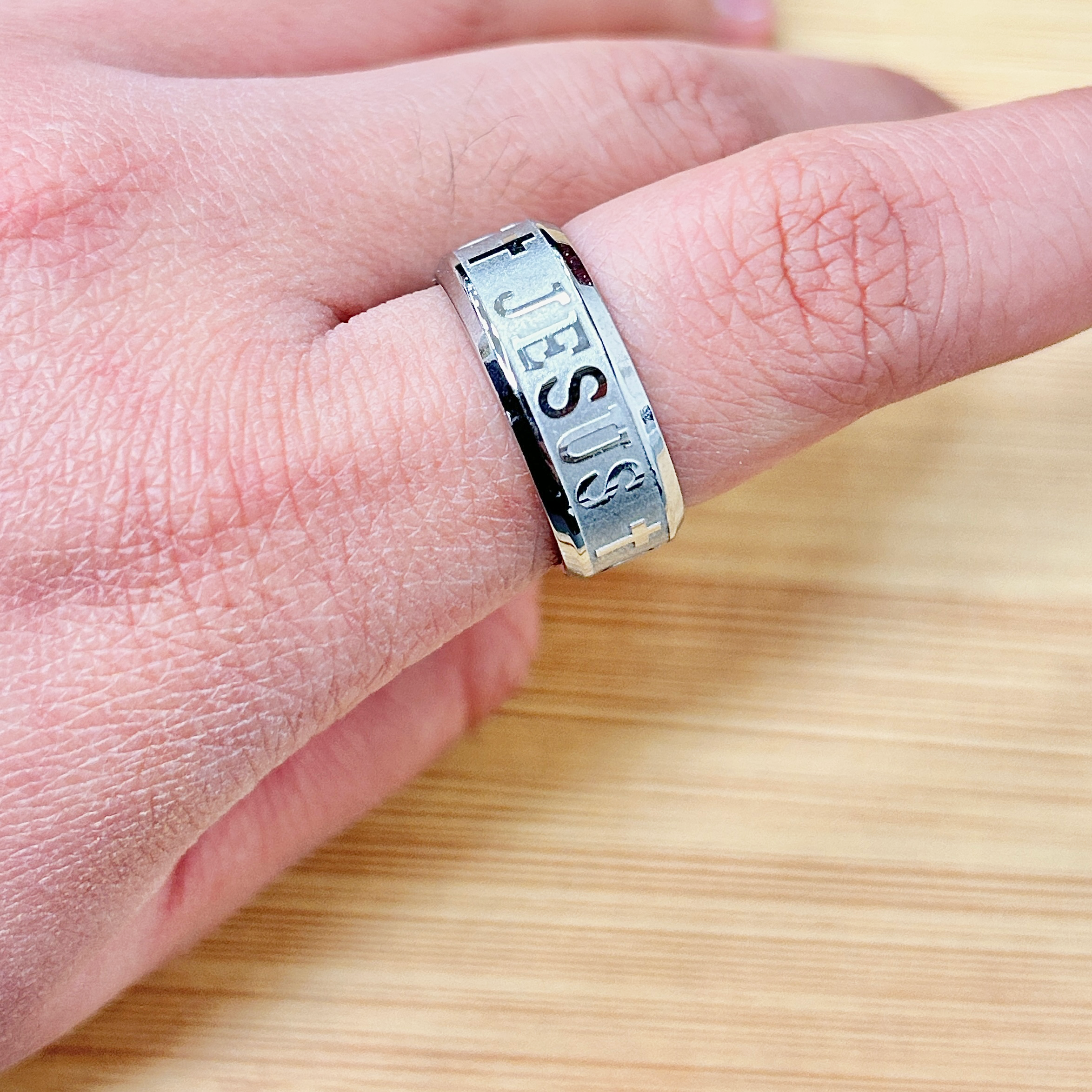 Silver Jesus Cross Band Ring - Eriy Gift Close-up of a silver ring with engraved letters on a finger over a wooden background.
