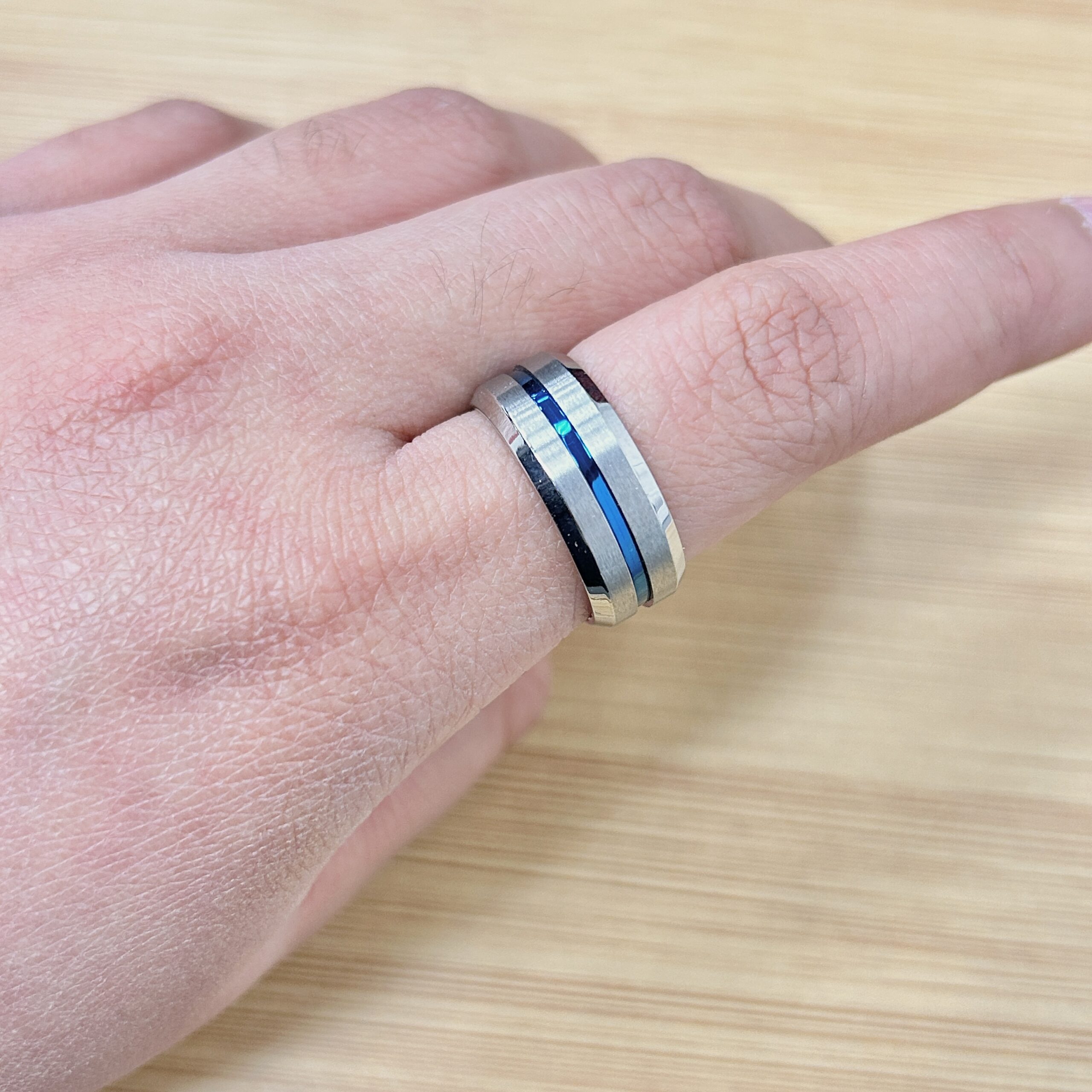 Close-up of a hand wearing a two-tone silver ring with a blue stripe, resting on a light wooden surface.