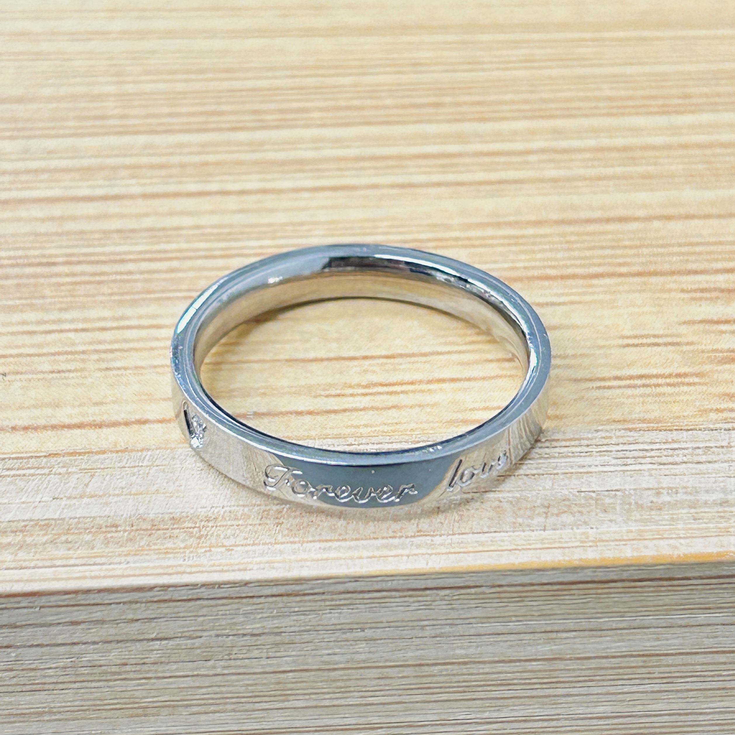 Silver Tone Forever Love Engraved Ring picture2 - Eriy Gift Close-up of a silver metal ring with 'Forever' engraved along the edge on a wooden surface.