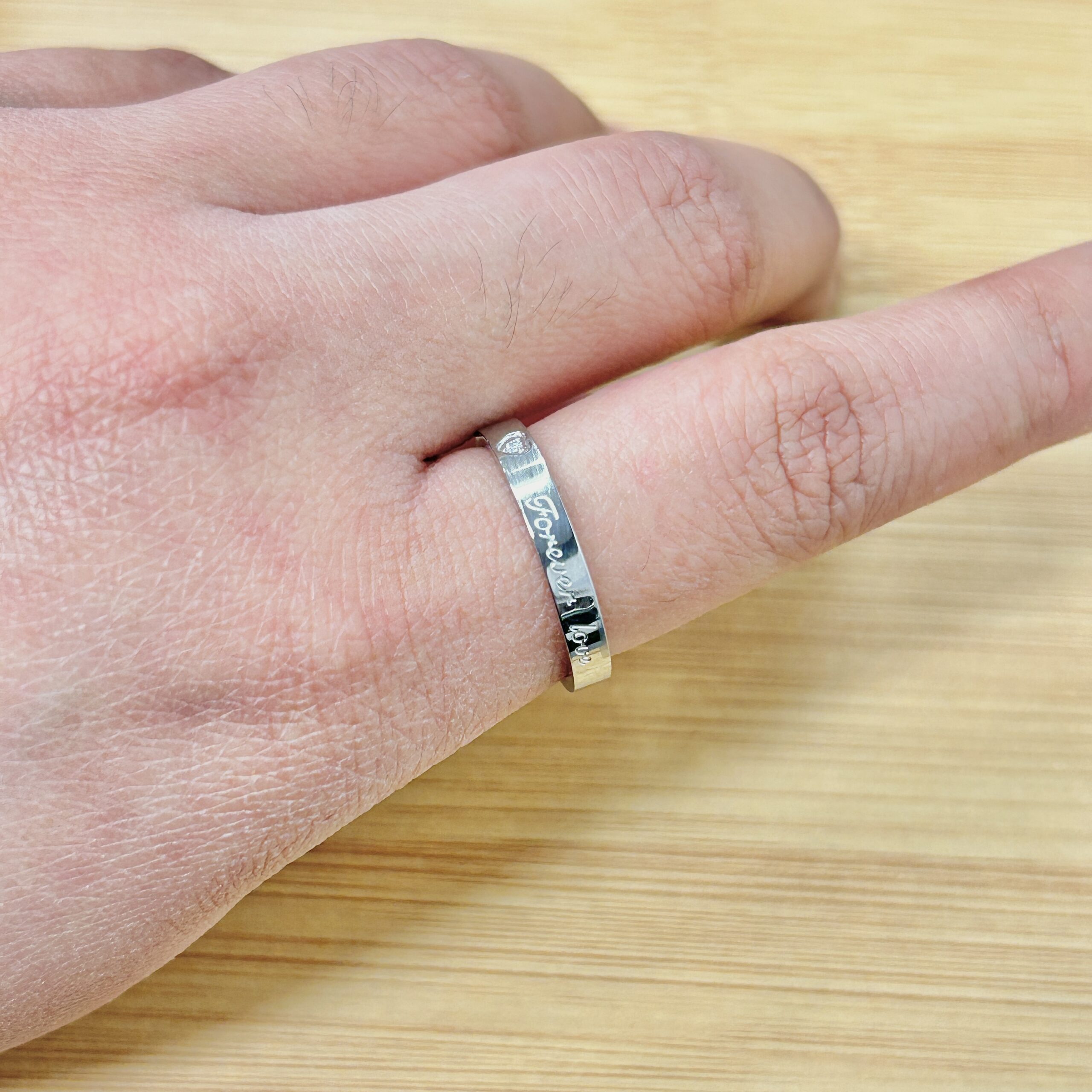 Close-up of a silver engraved ring on a finger resting on a light wood surface, with the word 'forever' visible on the band
