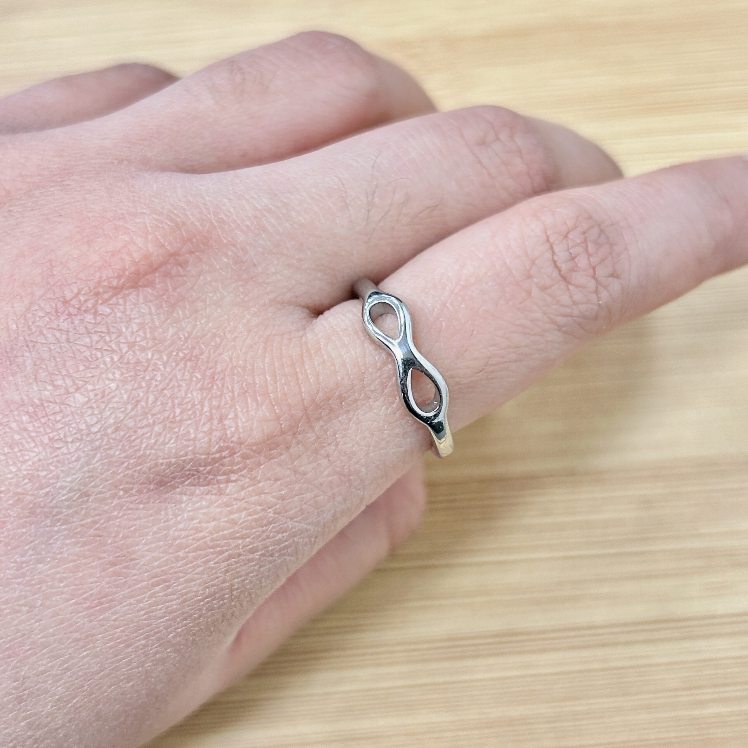 Close-up of a hand wearing a silver infinity-loop ring on the ring finger, resting on a light wooden surface.