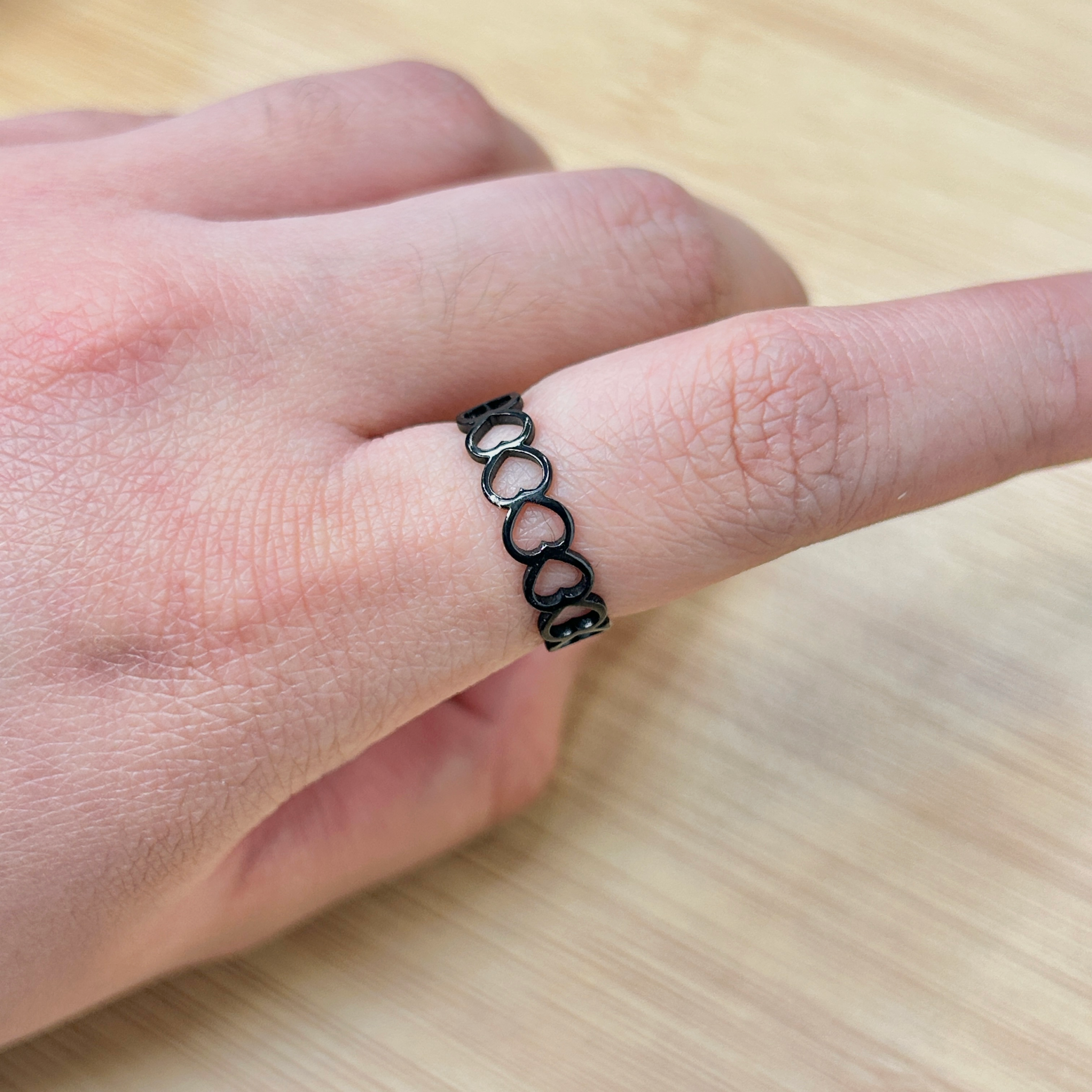 Close-up of a wrist with a black metal chain bracelet featuring connected heart-shaped links, resting on a light wooden surface.
