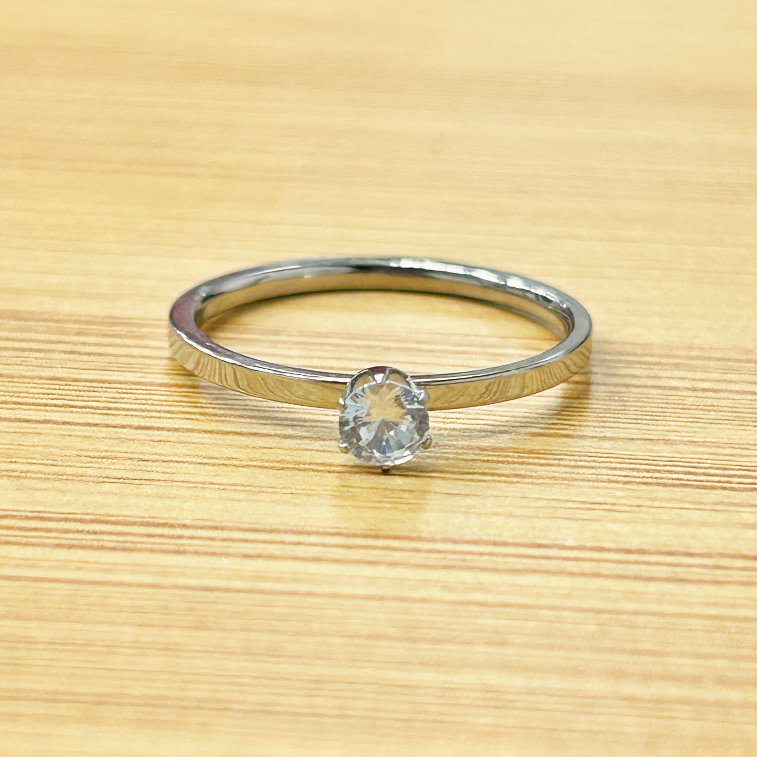 Thin Silver Solitaire Proposal Ring picture2 - Eriy Gift Close-up of a diamond engagement ring with a band resting on a light wooden surface, with the diamond facing the viewer.