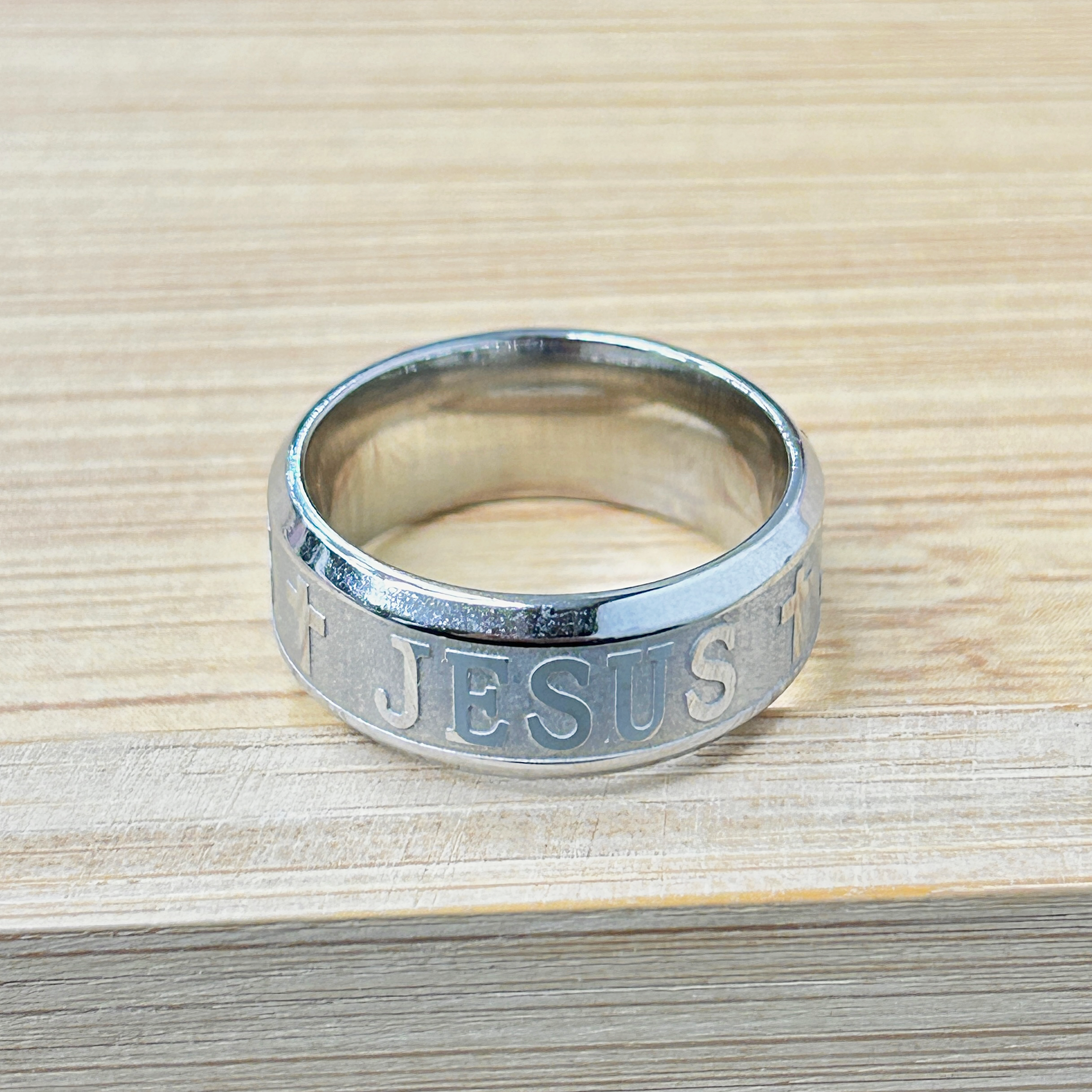 Vintage Silver Jesus Cross Band Ring picture2 - Eriy Gift Close-up of a metallic ring with the word 'JESUS' engraved along its side, resting on a light wood surface.