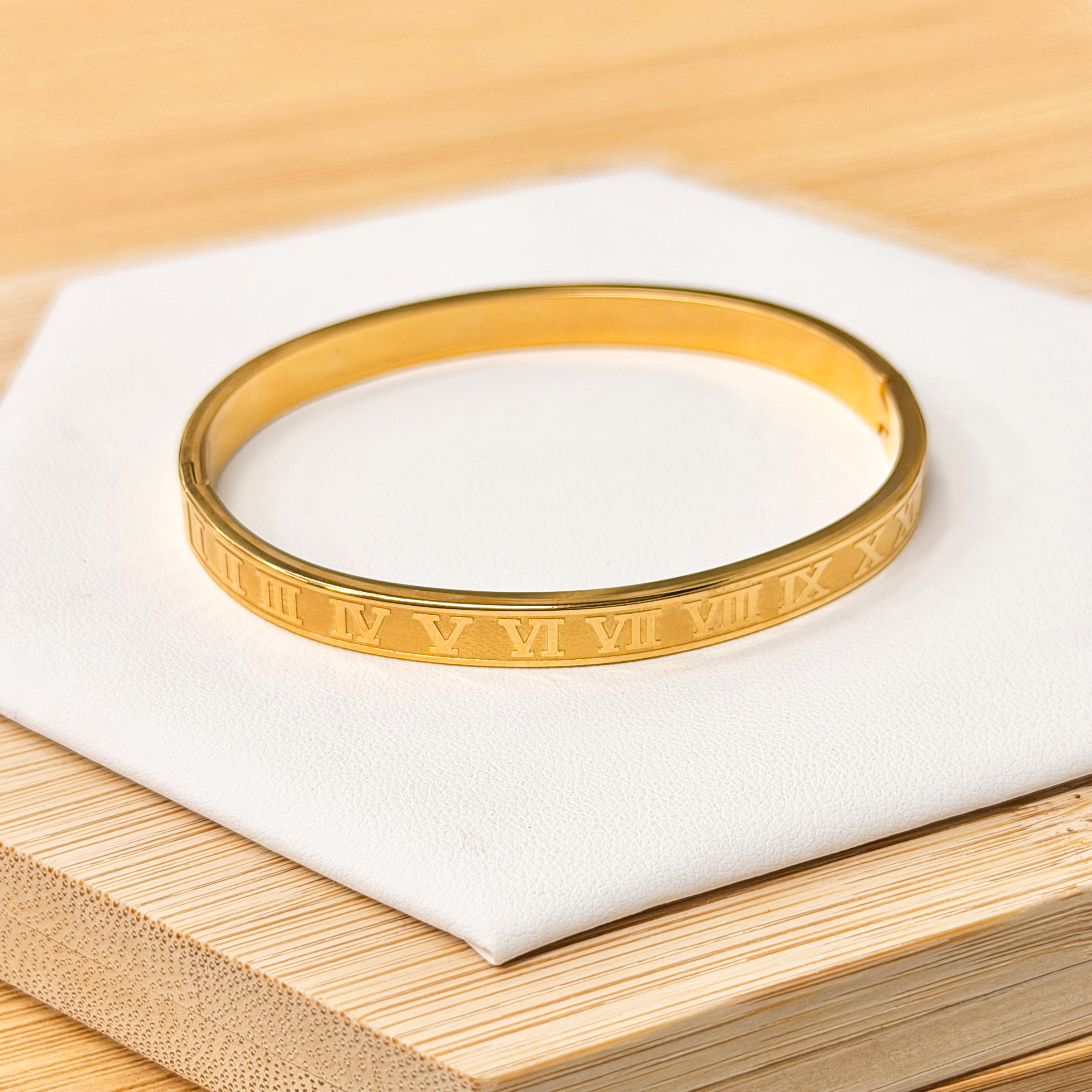 Classic Gold Embossed Roman Numeral Bangle picture1 - Eriy Gift Close-up of a gold bracelet (bangle) with engraved pattern resting on white fabric over a light wooden surface.