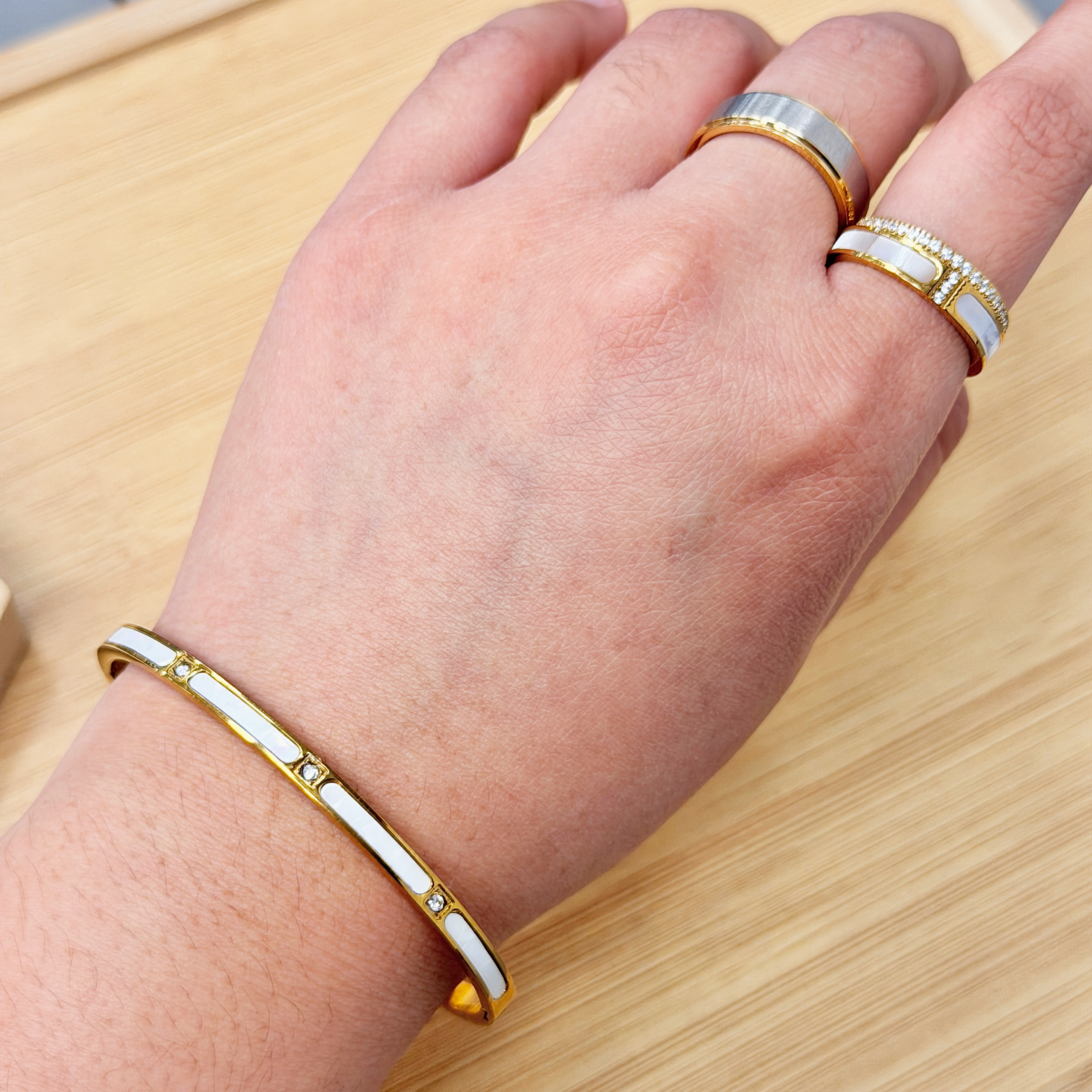 Elegant Gold Tone Opalescent Stone Inlay Bangle picture2 - Eriy Gift Close-up of a wrist wearing a gold and white enamel bracelet with diamonds on a light wooden surface.