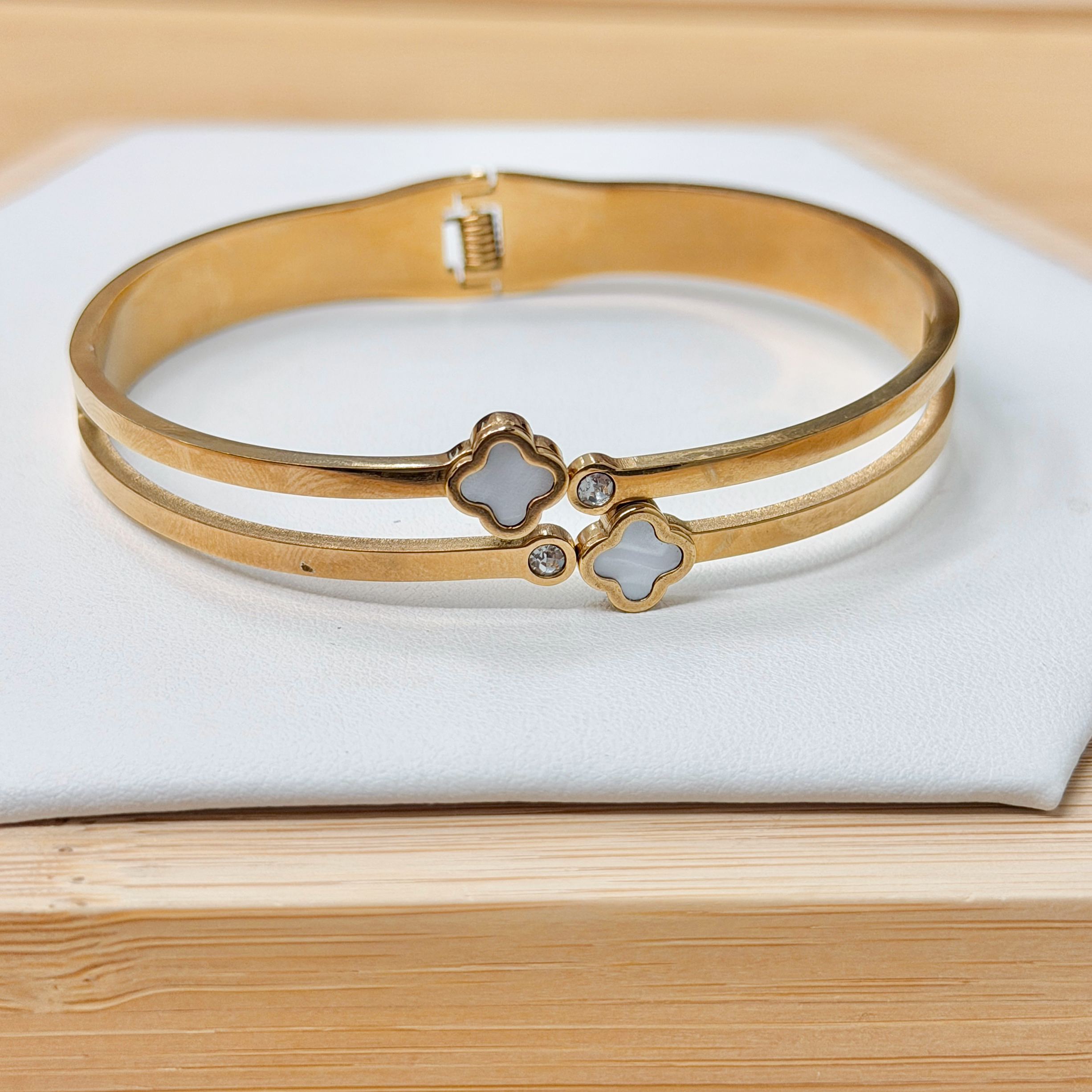 Gold Double Band Dual White Clover Bangle picture2 - Eriy Gift Gold-tone bracelet stack with clover-shaped charms resting on a white cushion atop a wooden surface