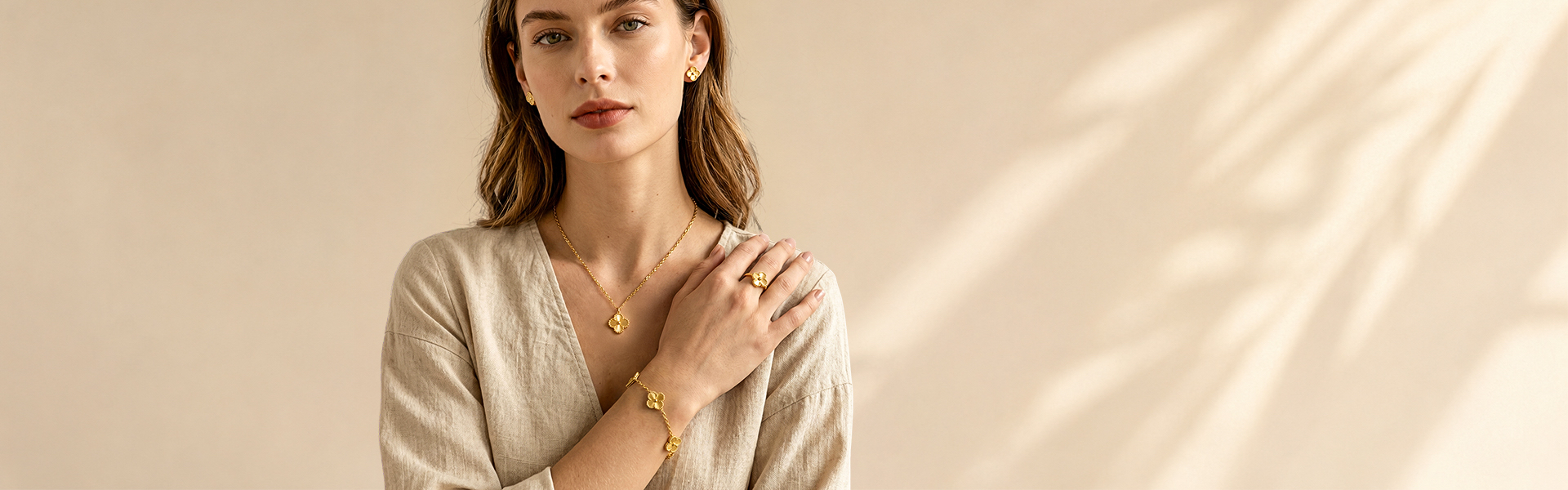Model wearing gold jewelry: necklace, bracelet, ring, and earrings on a light beige top with a warm beige backdrop.