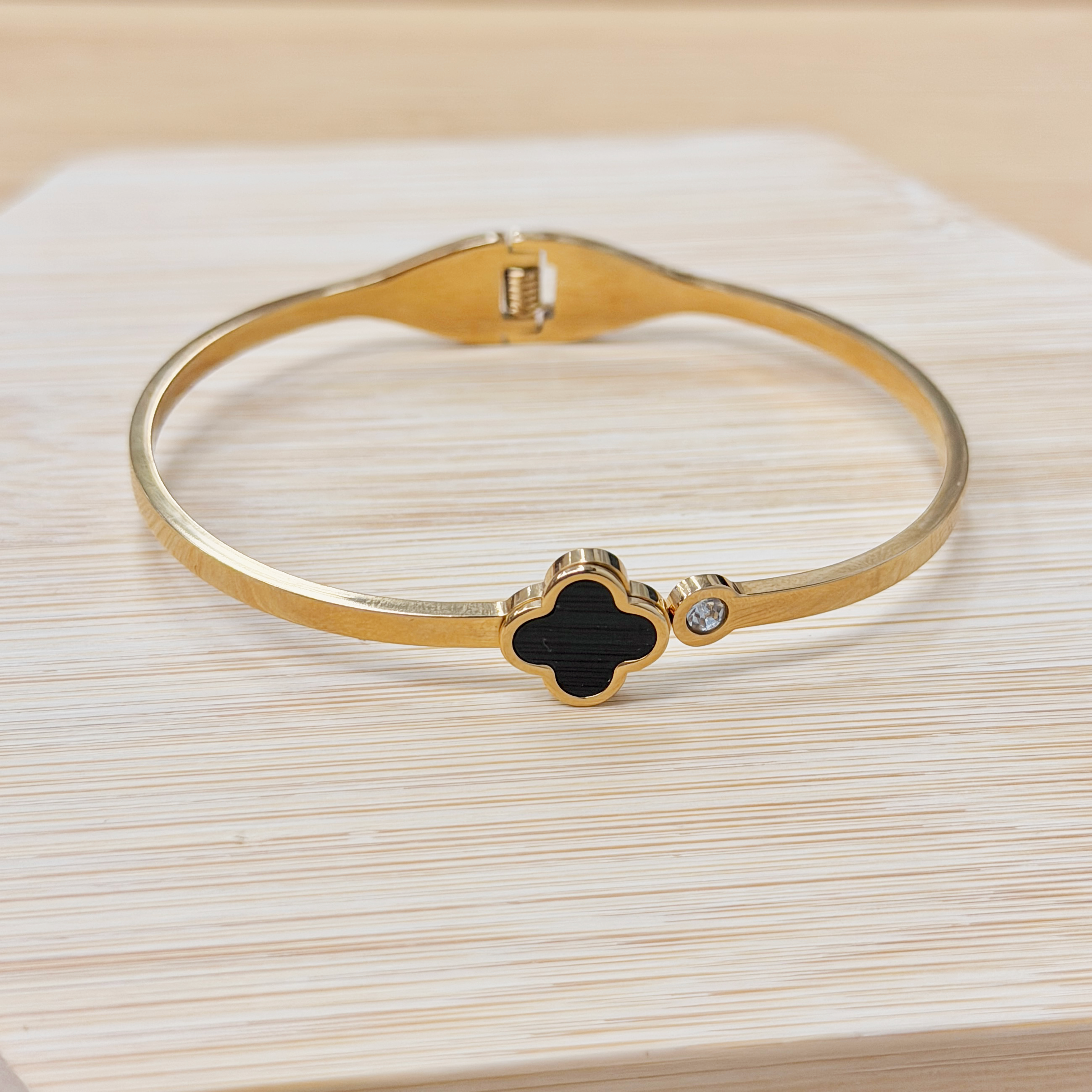 Close-up of a gold bangle bracelet with a black four-leaf-clover charm on a light wooden surface with visible grain