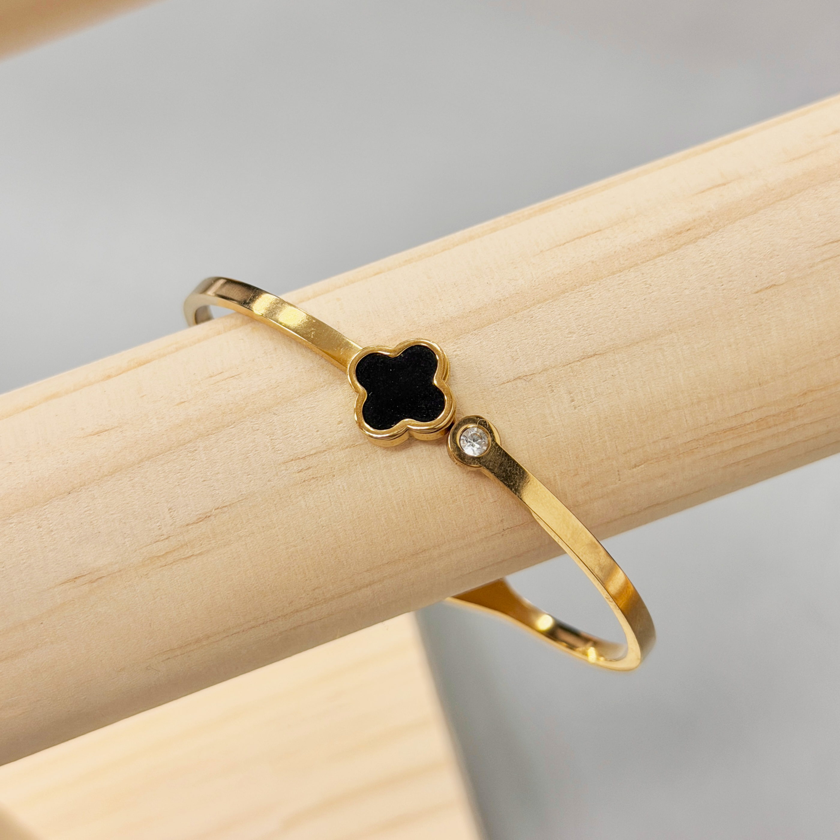 Gold bracelet with a clover-shaped black enamel charm resting on a light wooden surface.
