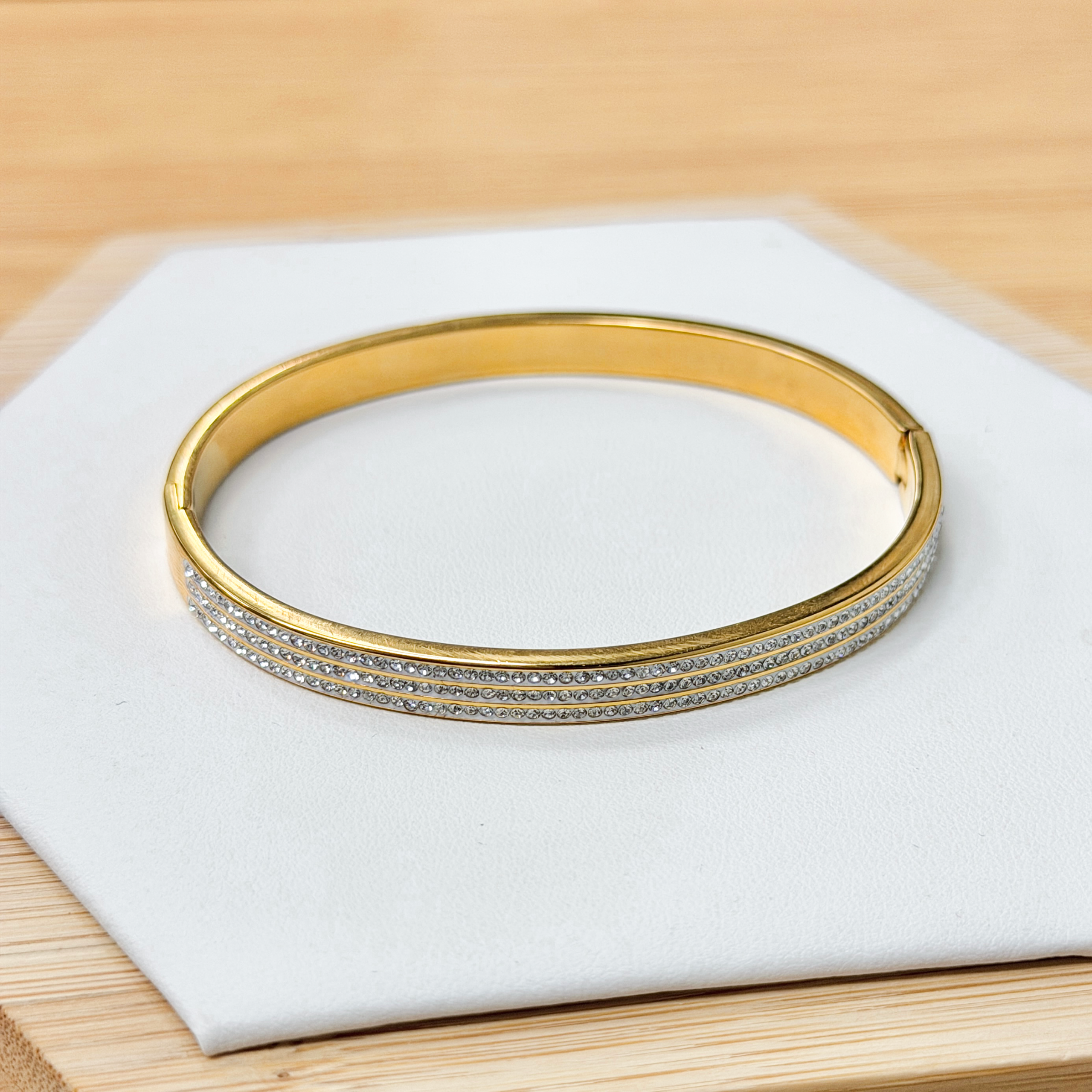 Zircon Paved Triple line Gold Bangle picture2 - Eriy Gift Gold bracelet with a row of diamonds, resting on a white display surface over a wooden table.